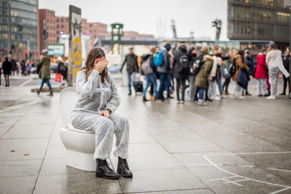 Eine Schülerin sitzt in einem weißen Overall auf einer Toilettenschüssel mitten auf dem Potsdamer Platz in Berlin.